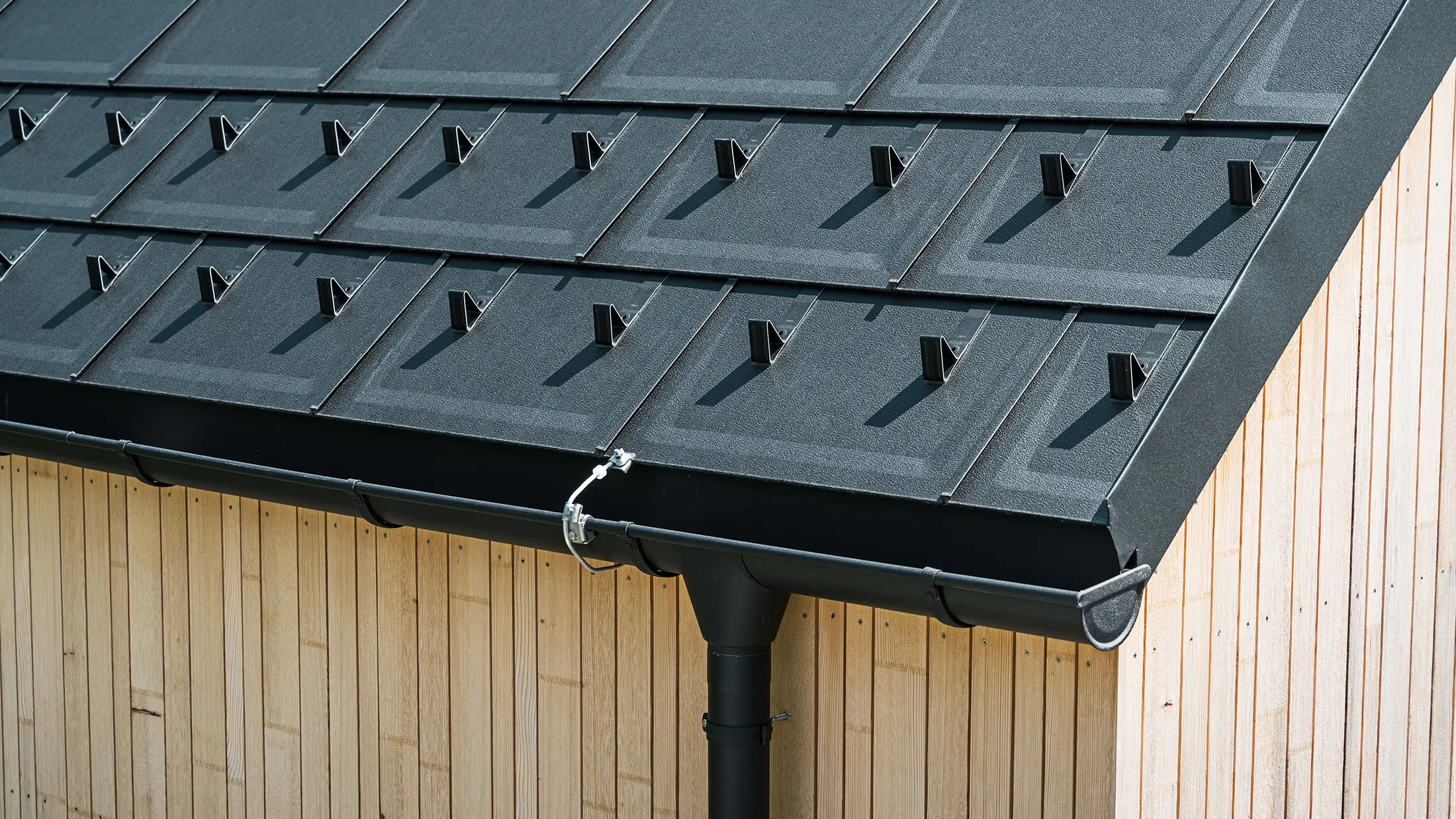 Prim plan al element classic PREFA R.16 cu cârlige parazăpadă, jgheab și burlan de culoare negru P.10 incl. paratrăsnet și o fațadă din lemn verticală. Produsele robuste din aluminiu PREFA împodobesc acoperișul în două ape al unei case monofamiliale din Lustenau, Vorarlberg.