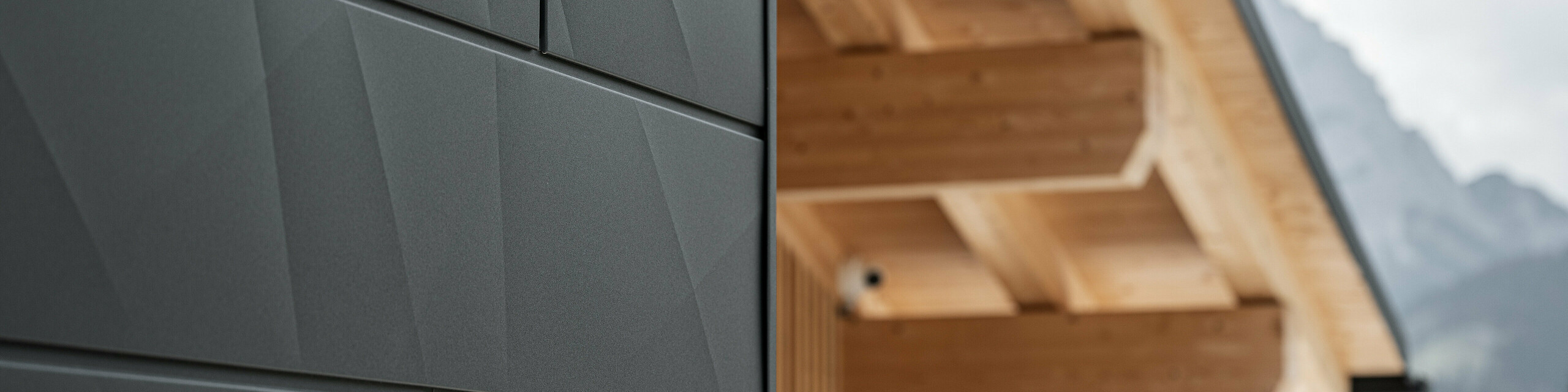Nahaufnahme der Siding.X Fassadenpaneele in P.10 Schwarz von PREFA an der Casa Hoffe in Sappada, Italien. Das Fassadensystem aus Aluminium ist gekonnt in einem modernen, abstrakten Design angebracht, das einen ansprechenden Kontrast zu den sichtbaren Holzelementen des Dachstuhls bildet. Die innovative Fassadenlösung fügt sich nahtlos in das alpine Ambiente ein und steht für die Verbindung von Funktionalität, Beständigkeit und ästhetischer Baukultur.