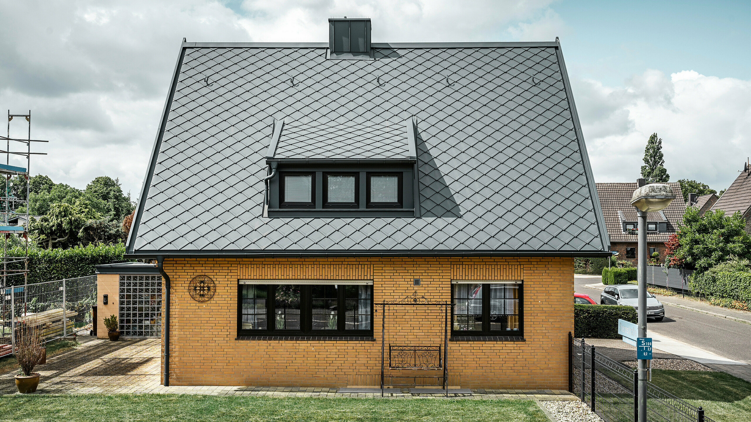 Seitenansicht eines Einfamilienhauses in Tönisvorst mit einem anthrazitfarbenen PREFA Dach aus Dachrauten 29×29. Die Kombination des traditionellen gelben Backsteins mit den modernen Dachelementen und Zubehör aus Aluminium ergibt ein harmonisches Gesamtbild. Schleppgauben und Kamin, ebenfalls in Anthrazit gehalten, vervollständigen das ästhetische und funktionale Dach in einer kleinstädtischen Umgebung.