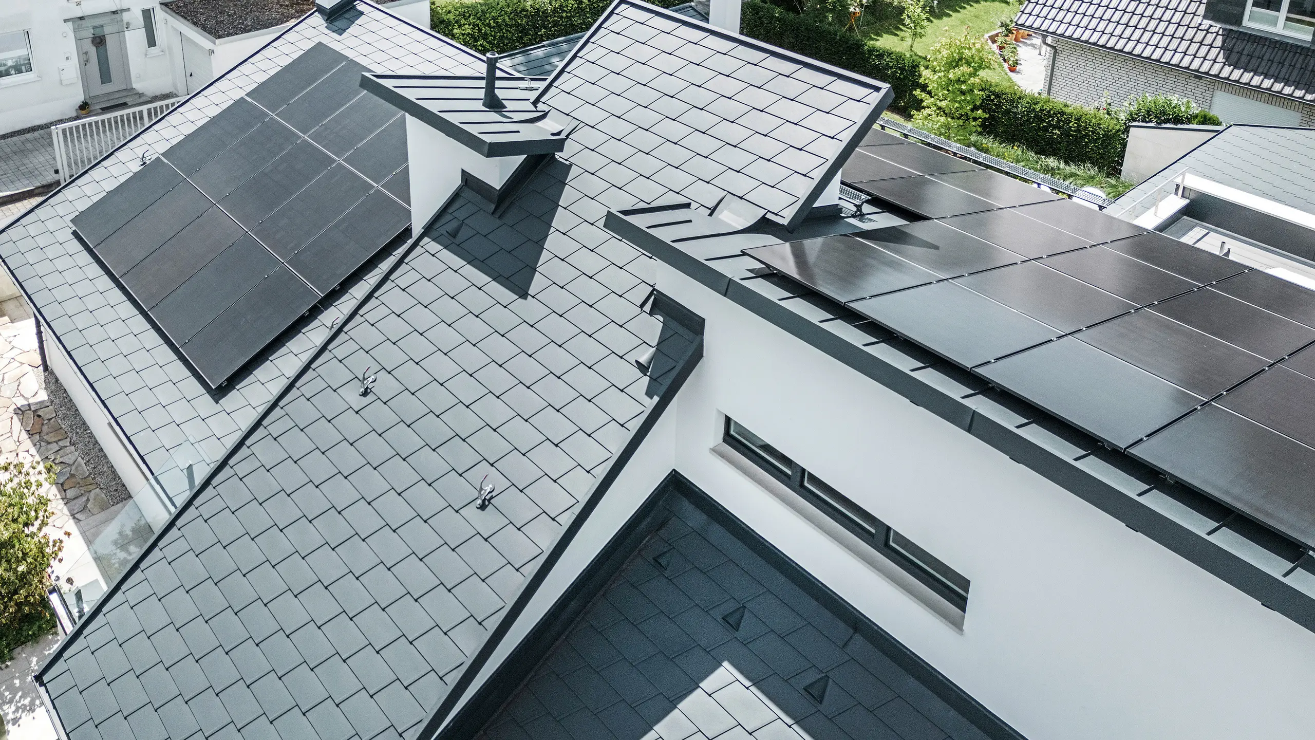 Casă unifamilială modernă din Dortmund, cu o instalație fotovoltaică avansată, montată pe un sistem de acoperiș PREFA alcătuit din șindrile DS.19 și o învelitoare cu falț vertical PREFALZ în culoarea P.10 antracit. Instalația fotovoltaică este fixată cu ajutorul suportului Vario pentru panouri solare destinat șindrilelor și al suportului PREFALZ Vario pentru panouri solare destinat benzilor cu falț vertical. Instalația solară asigură producția eficientă a energiei din surse regenerabile. Designul bine conceput al acoperișului oferă un echilibru optim între estetică și funcționalitate.