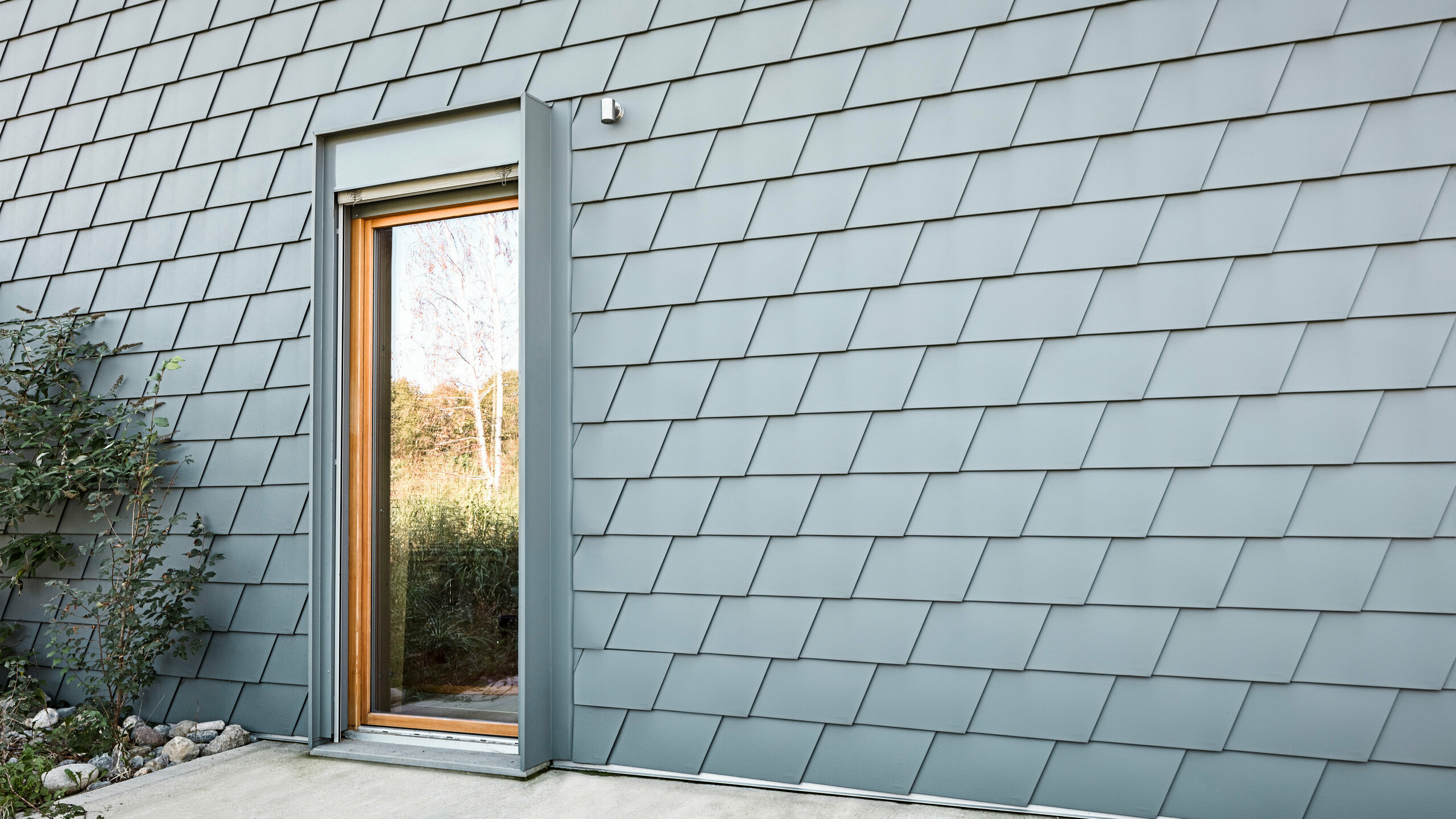 Fassadenansicht des Casa Longaretti mit PREFA Dachschindeln in P.10 Hellgrau, die eine schuppenartige Textur bilden und dem Gebäude einen modernen Charakter verleihen. Die harmonische Farbgebung und die präzise Verlegung der Schindeln unterstreichen die Premiumqualität und das innovative Design von PREFA Aluminiumprodukten. Der Holzrahmen der Tür fügt einen natürlichen Kontrast hinzu, der das ästhetische und nachhaltige Konzept dieses Objekts hervorhebt.