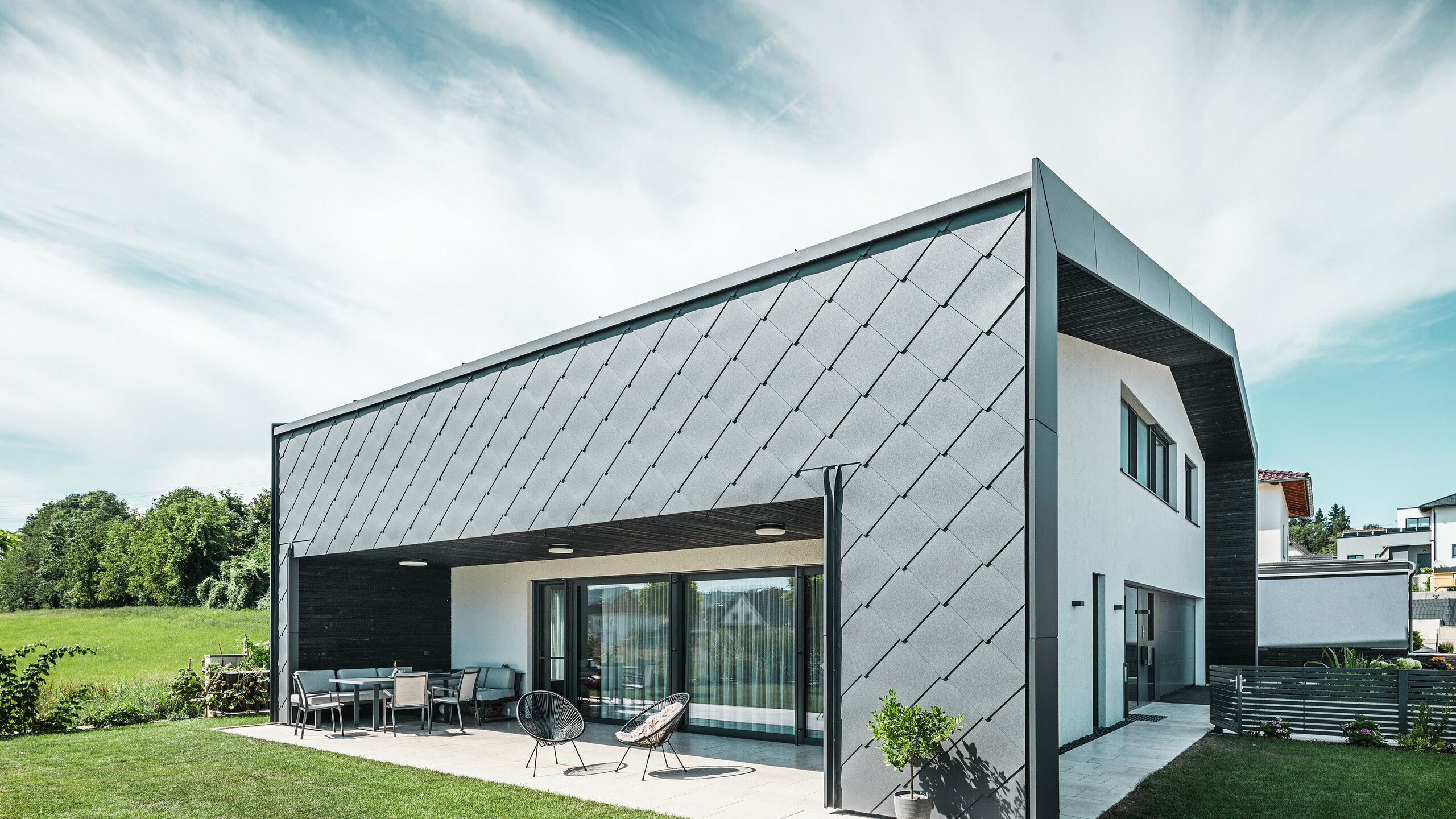 Grădină îngrijită a unei case unifamiliale din Aschach. Clădirea este acoperită elegant cu panouri PREFA din aluminiu de culoare gri închis, cu dimensiuni de 44 × 44. Terasa acoperită oferă un loc confortabil pentru relaxare, cu vedere la peluza verde și luxuriantă. Acoperirea elegantă din aluminiu, în combinație cu suprafețele tencuite în alb, asigură o arhitectură modernă și rezistentă la intemperii. Ferestrele mari permit pătrunderea luminii naturale în interior și creează o atmosferă deschisă și luminoasă.