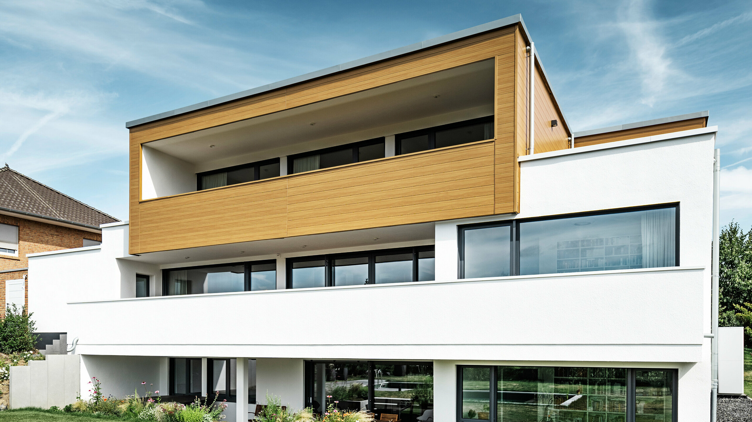 Die Rückseite eines modernen Einfamilienhauses in Usingen, Deutschland. Am unteren Bildrand ist die grüne Wiese, ein Pool sowie ein junger Gartenbaum zu sehen. Das auffällige Haus verfügt über einen weißen Gebäudesockel aus einer Putzfassade, einer Aluminiumfassade am Obergeschoss aus PREFA Sidings in Eiche natur sowie großen Fensterflächen mit einem anthrazitfarbenen Rahmen. Aus der Distanz ist das Nachbarhaus mit einer Ziegelfassade am linken Bildrand zu erkennen.