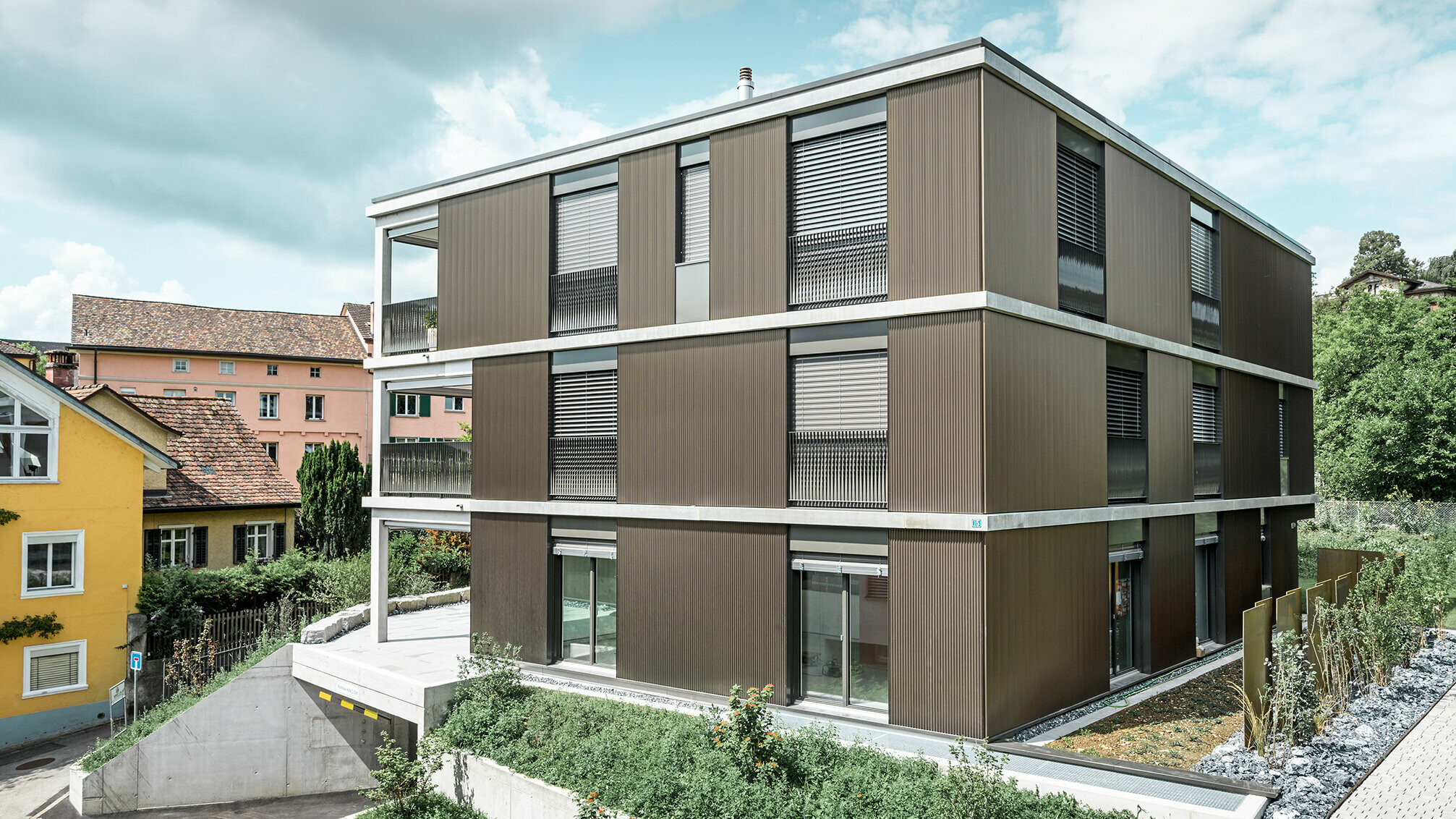 Die markante Aluminiumfassade dieses Mehrfamilienhauses in Schaffhausen ist mit dem eleganten PREFA Zackenprofil in der Farbe Colinal ausgestattet. Durch die vertikale Anordnung der stranggepressten Aluminiumprofile wirkt das Gebäude optisch kompakter und die Fassadenstruktur erhält eine harmonische Balance. Das edle Design fügt sich geschmackvoll in die Umgebung ein und verleiht dem Gebäude eine moderne Eleganz, die sowohl optisch ansprechend als auch funktional ist. Die Perspektive zeigt das Mehrfamilienhaus von der Vorderseite mit der Garageneinfahrt. Im Hintergrund sind Bäume, weitere Gebäude und ein Kirchturm zu erkennen.