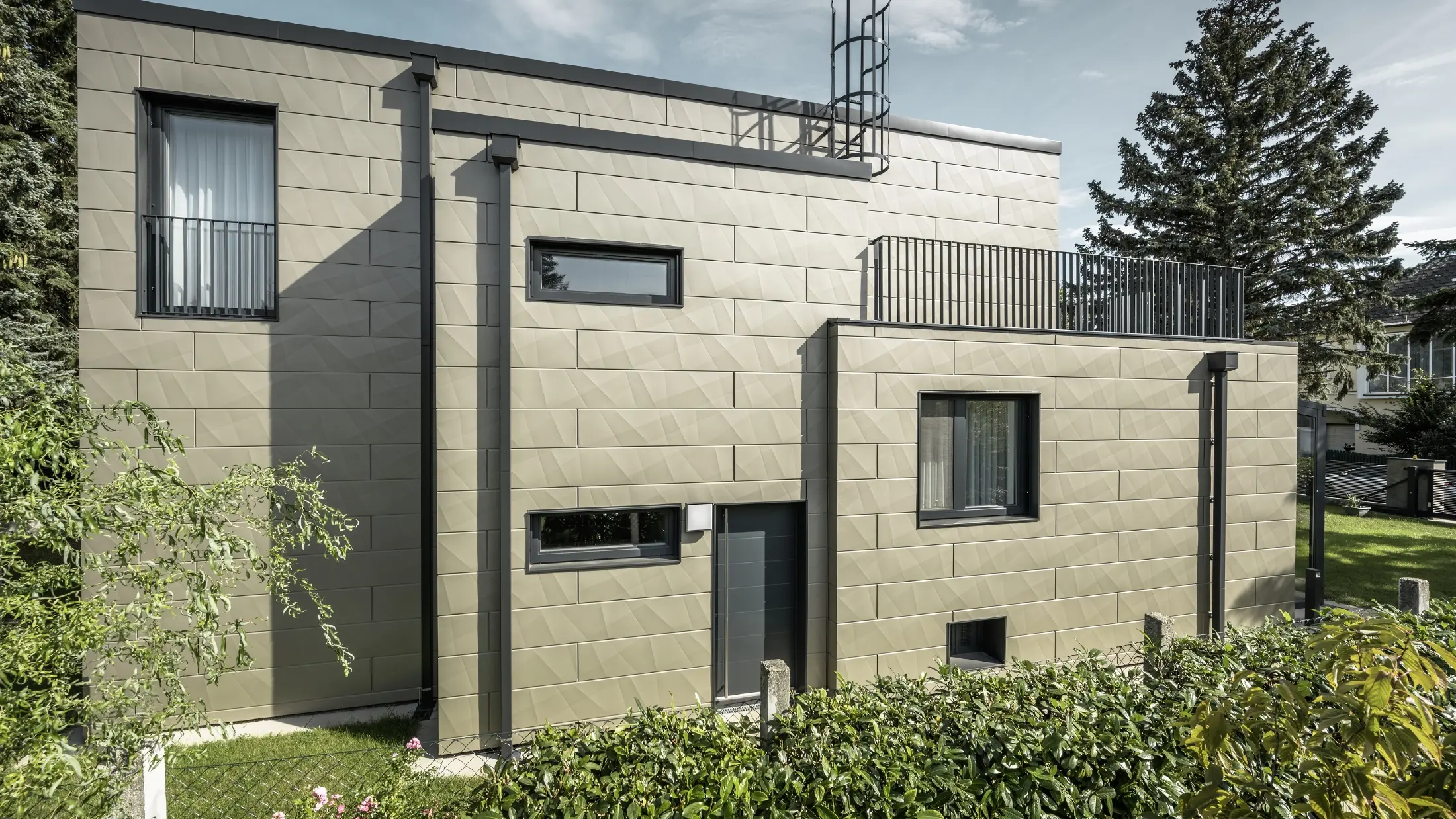 Construcție nouă cu acoperiș plat, fațada este placată cu panoul pentru fațade Siding.X PREFA cu ambutisări în culoarea bronz, ancadramentele, precum și burlanul rectangular, burlanul dreptunghiular PREFA sunt realizate în culoarea antracit.,   