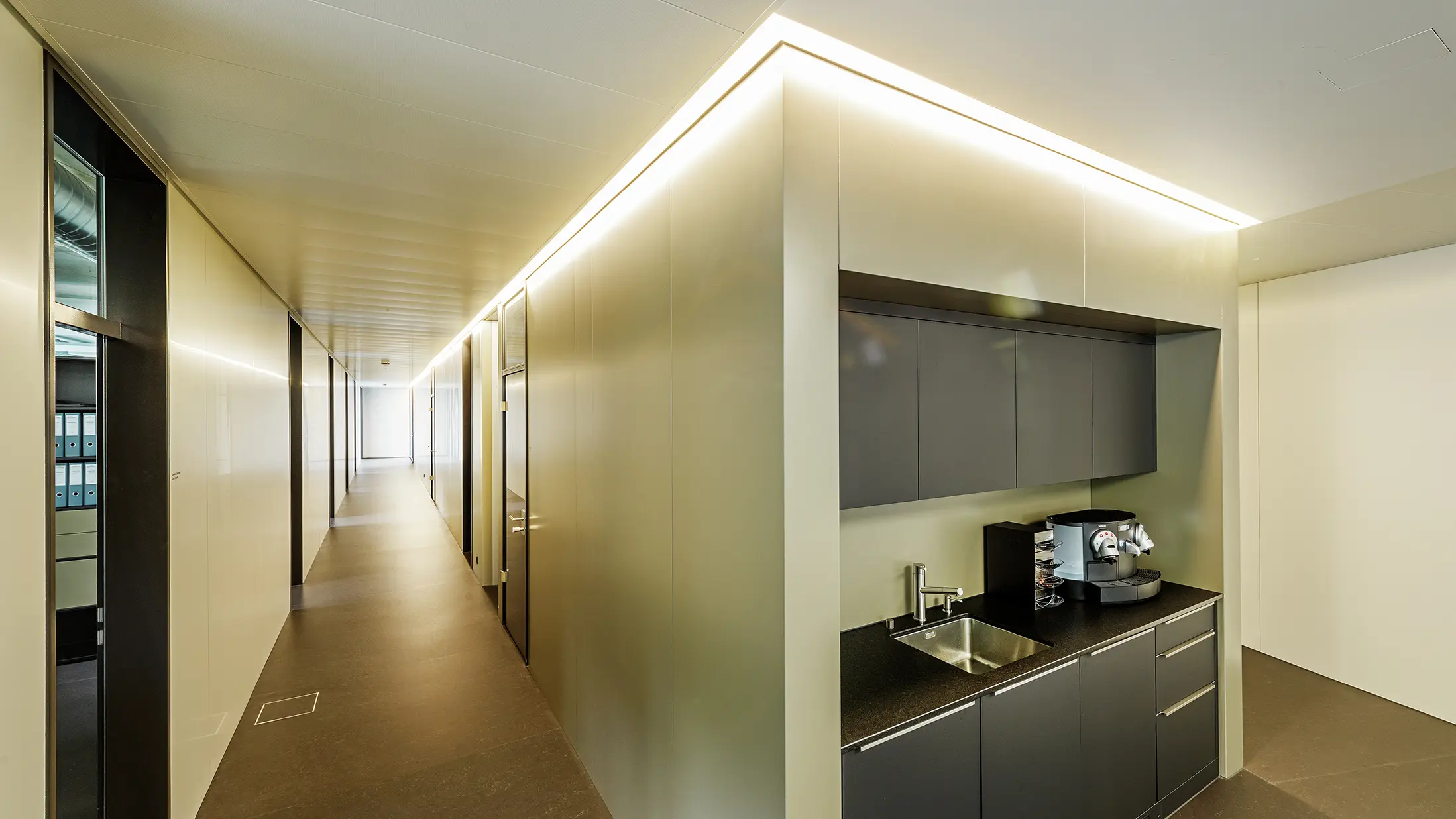 În spațiile moderne de birouri ale firmei Guggisberg Dachtechnik din Wabern, chicineta integrată asigură funcționalitate și design. Placările de perete preiau aspectul elegant de bronz al fațadei din aluminiu PREFA și se integrează perfect în conceptul arhitectural. Amenajarea interioară cu iluminare indirectă creează o senzație plăcută de spațiu.