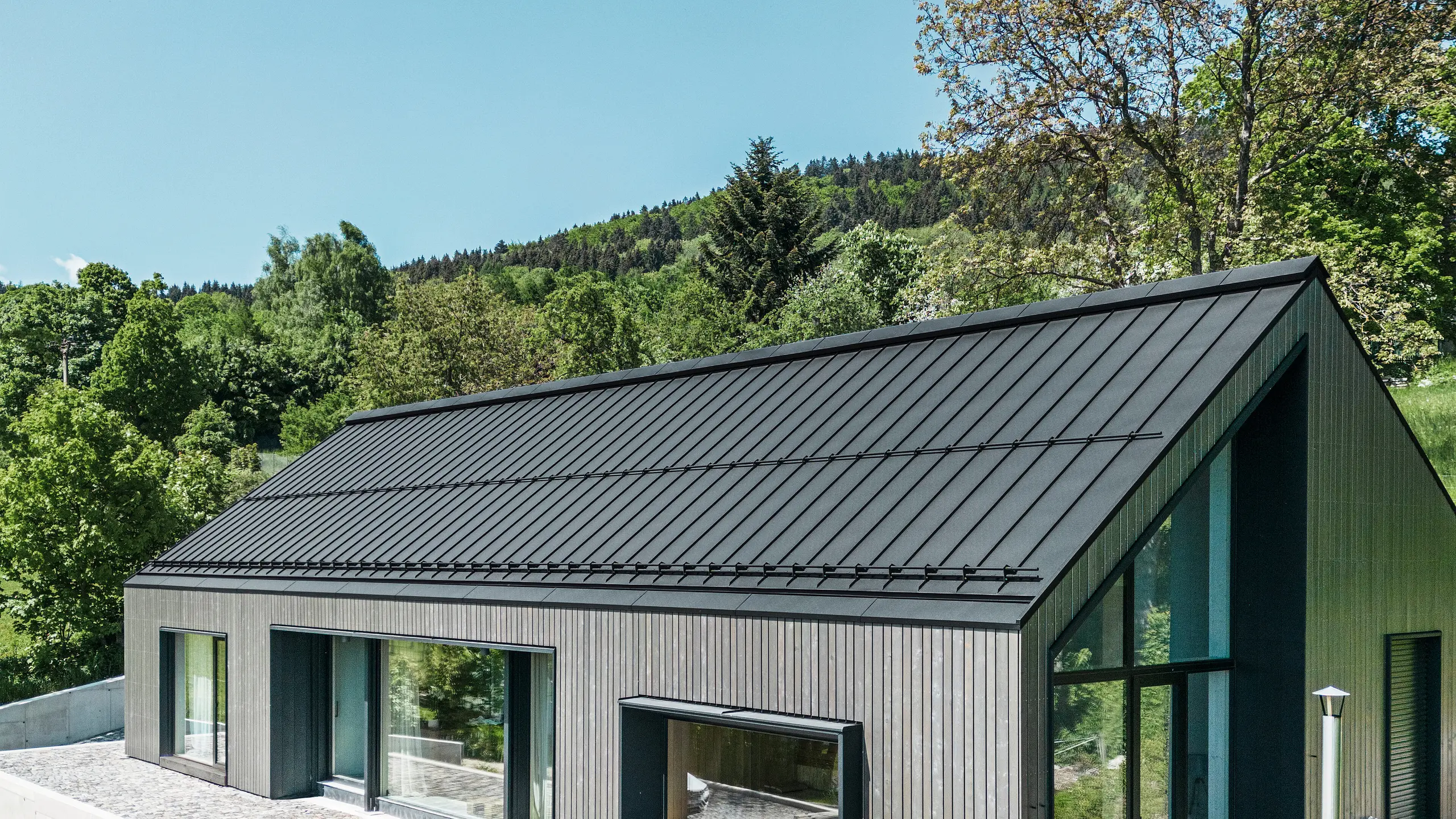Ein modernes Holzmassivhaus in Kuncice (CZ) wurde mit großen Fensterflächen ausgestattet. Das dunkle Holzmassivhaus verfügt über ein Dachsystem aus PREFALZ in der Farbe P. 10 Schwarz. Im hinteren Außenbereich steht ein Whirlpool mit einer Holzverkleidung.
