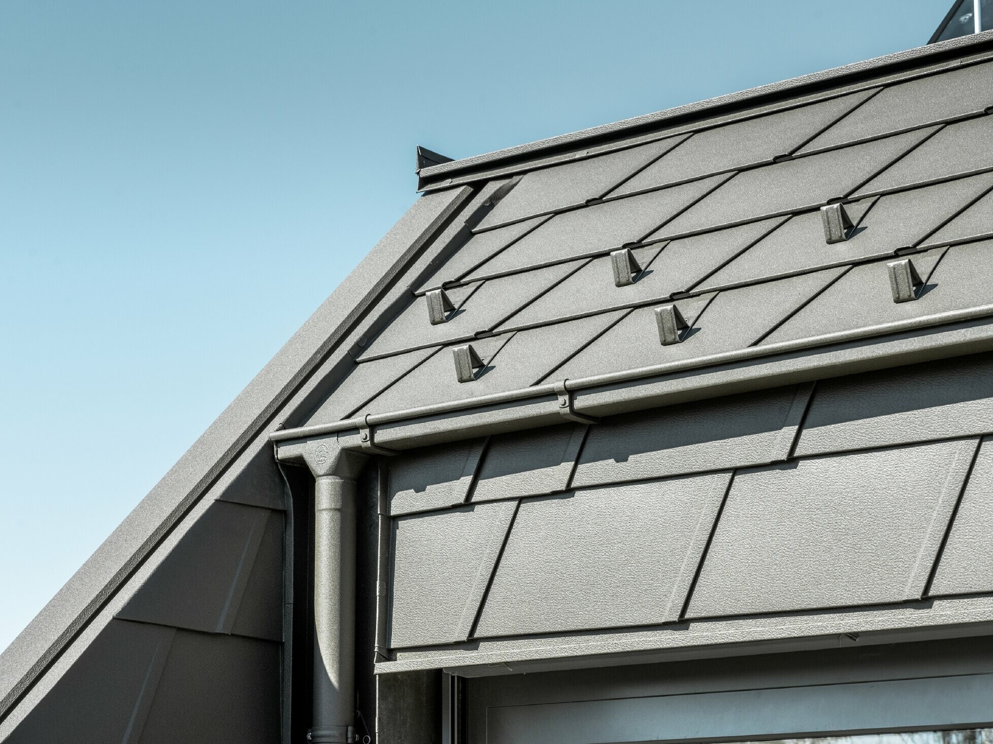 Șindrile de aluminiu PREFA DS.19 cu opritor de zăpadă. Drenajul acoperișului se realizează prin intermediul țevii pătrate PREFA și al țevii de scurgere în culoarea P.10 maro.