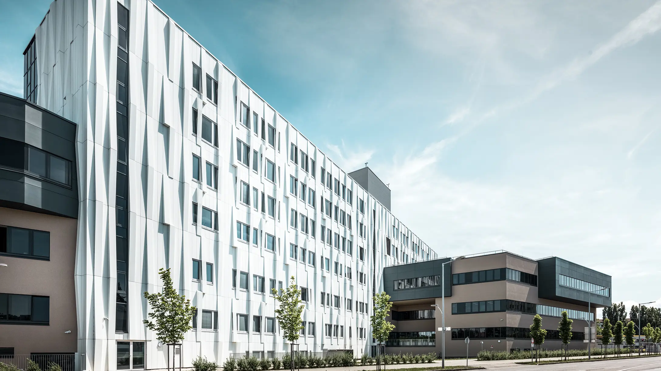 Seitliche Ansicht des Universitätsklinikums in St. Pölten, Niederösterreich. Die robuste Aluminiumfassade macht das Gebäude zu einem Blickfang in der Stadt. Im Vordergrund ist eine Straße mit einigen Bäumen zu sehen.