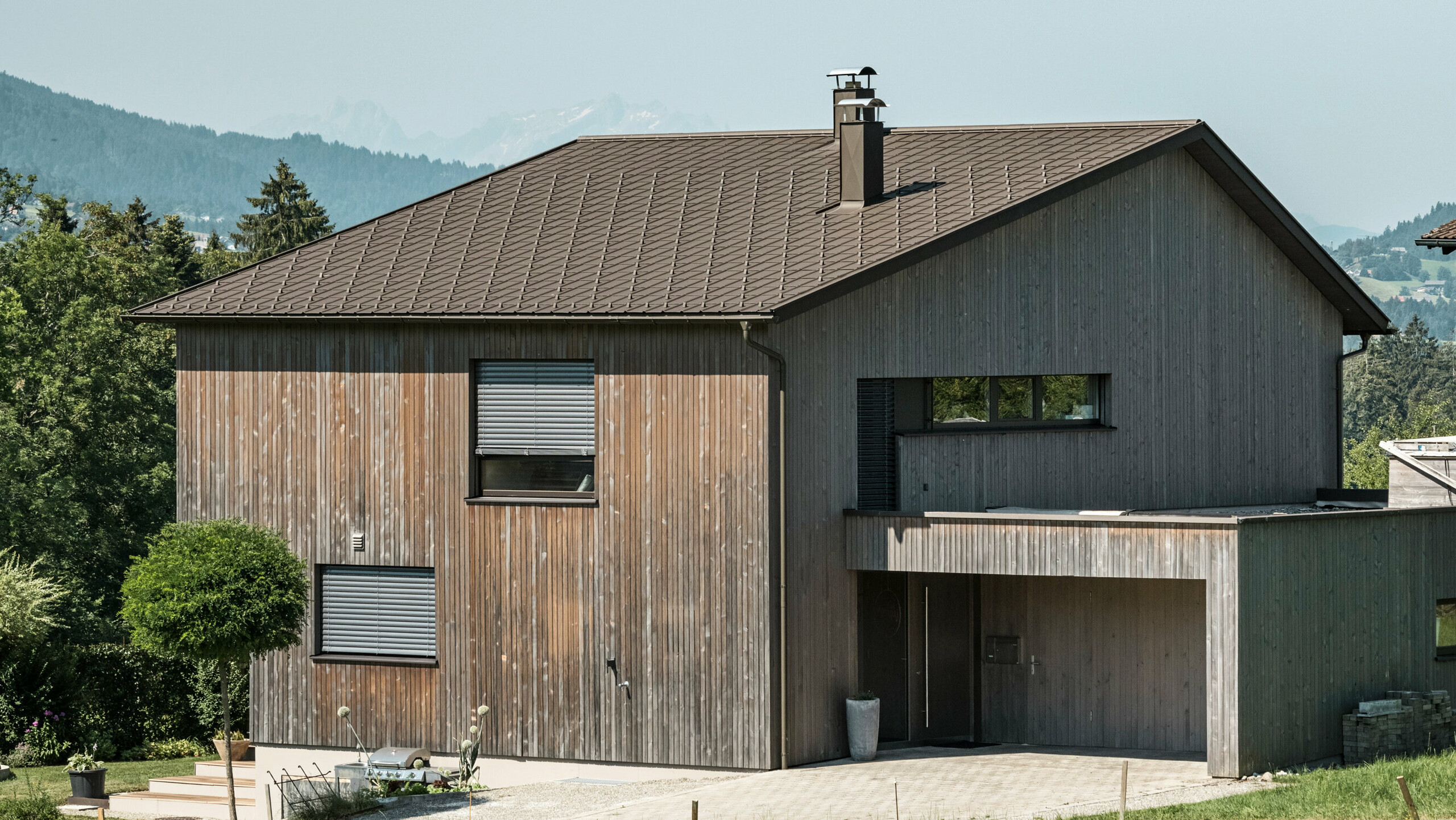 Seitenansicht eines zeitgenössischen Einfamilienhauses in Krumbach mit einer Kombination aus einer natürlichen Holzfassade und einem langlebigen PREFA Aluminiumdach in P.10 Braun. Die harmonische Farbgebung zeigt die elegante Integration von modernem Design und traditionellen Materialien, wobei die PREFA Dachplatten für eine ästhetische und funktionale Dachgestaltung sorgen.