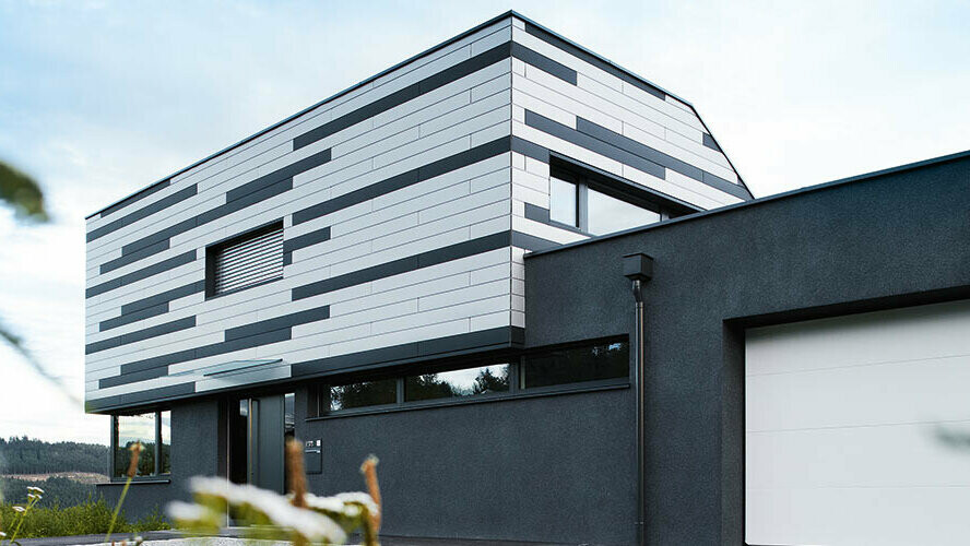 Das Bild zeigt die moderne Fassade eines Einfamilienhauses, das mit PREFA Sidings in den Farben Silber und Anthrazit matt gestaltet wurde. Die Platten sind in einem dynamischen, horizontalen Muster verlegt, wodurch der Architektur des Hauses ein stilvolles, zeitgemäßes Erscheinungsbild verliehen wird. Die Kombination aus den schlichten Farben und der klaren Linienführung sorgt für einen eleganten, aber zugleich markanten Look.