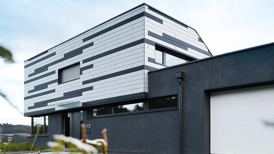 Das Bild zeigt die moderne Fassade eines Einfamilienhauses, das mit PREFA Sidings in den Farben Silber und Anthrazit matt gestaltet wurde. Die Platten sind in einem dynamischen, horizontalen Muster verlegt, wodurch der Architektur des Hauses ein stilvolles, zeitgemäßes Erscheinungsbild verliehen wird. Die Kombination aus den schlichten Farben und der klaren Linienführung sorgt für einen eleganten, aber zugleich markanten Look.