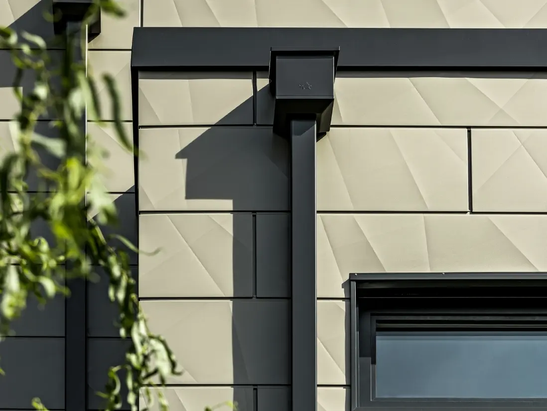 Casă monofamilială modernă cu acoperiș plat și geamuri înguste. Fațada este placată cu PREFA Siding.X cu aspect încrețit în culoarea bronz. Conectările au fost realizate în culoarea antracit. Ca sistem pluvial s-a folosit burlanul dreptunghiular PREFA cu căminul colector de apă potrivit, în culoarea antracit.
