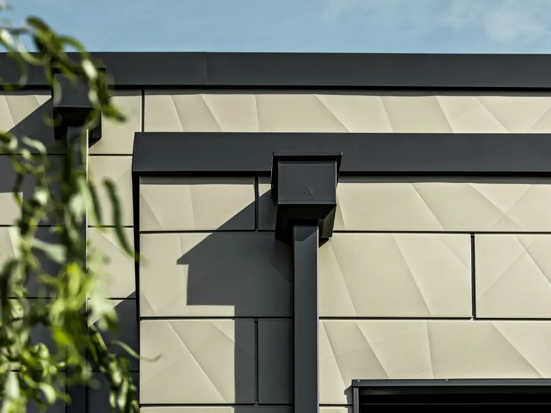Casă monofamilială modernă cu acoperiș plat și geamuri înguste. Fațada este placată cu PREFA Siding.X cu aspect încrețit în culoarea bronz. Conectările au fost realizate în culoarea antracit. Ca sistem pluvial s-a folosit burlanul dreptunghiular PREFA cu căminul colector de apă potrivit, în culoarea antracit.