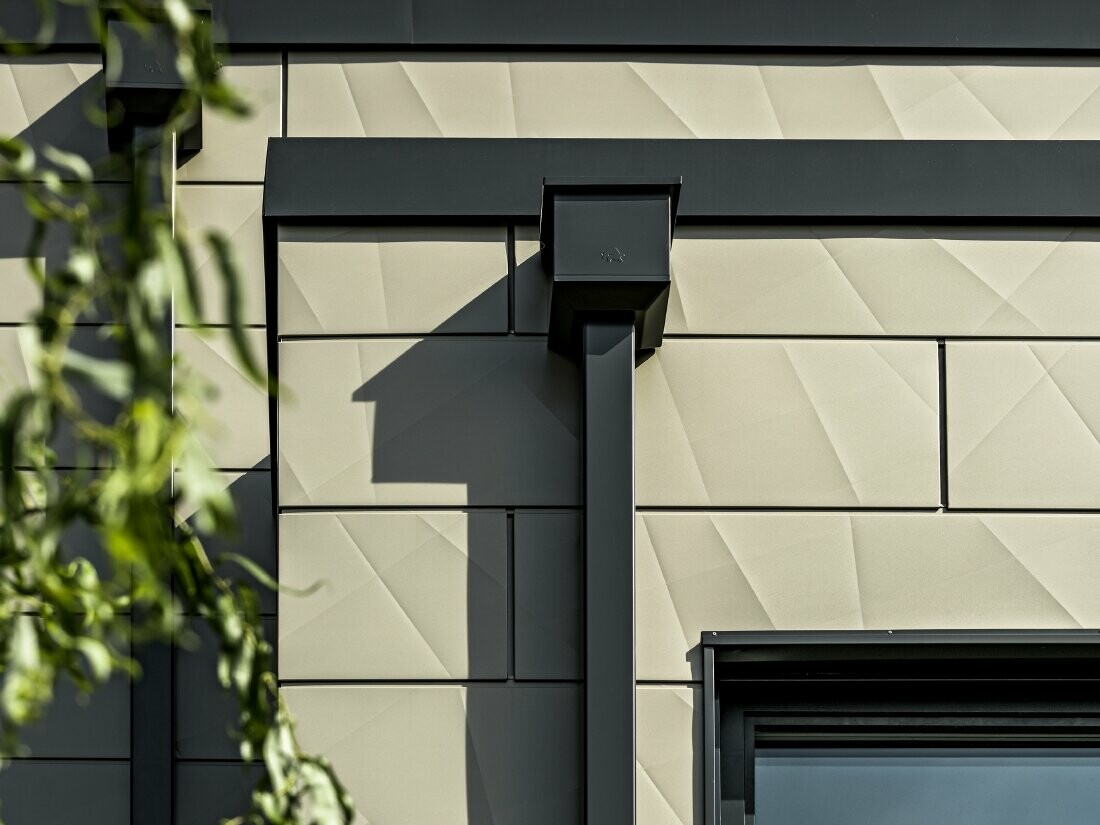 Casă monofamilială modernă cu acoperiș plat și geamuri înguste. Fațada este placată cu PREFA Siding.X cu aspect încrețit în culoarea bronz. Conectările au fost realizate în culoarea antracit. Ca sistem pluvial s-a folosit burlanul dreptunghiular PREFA cu căminul colector de apă potrivit, în culoarea antracit.