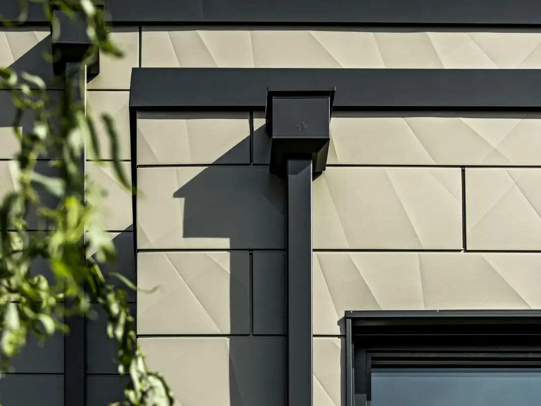 Casă monofamilială modernă cu acoperiș plat și geamuri înguste. Fațada este placată cu PREFA Siding.X cu aspect încrețit în culoarea bronz. Conectările au fost realizate în culoarea antracit. Ca sistem pluvial s-a folosit burlanul dreptunghiular PREFA cu căminul colector de apă potrivit, în culoarea antracit.