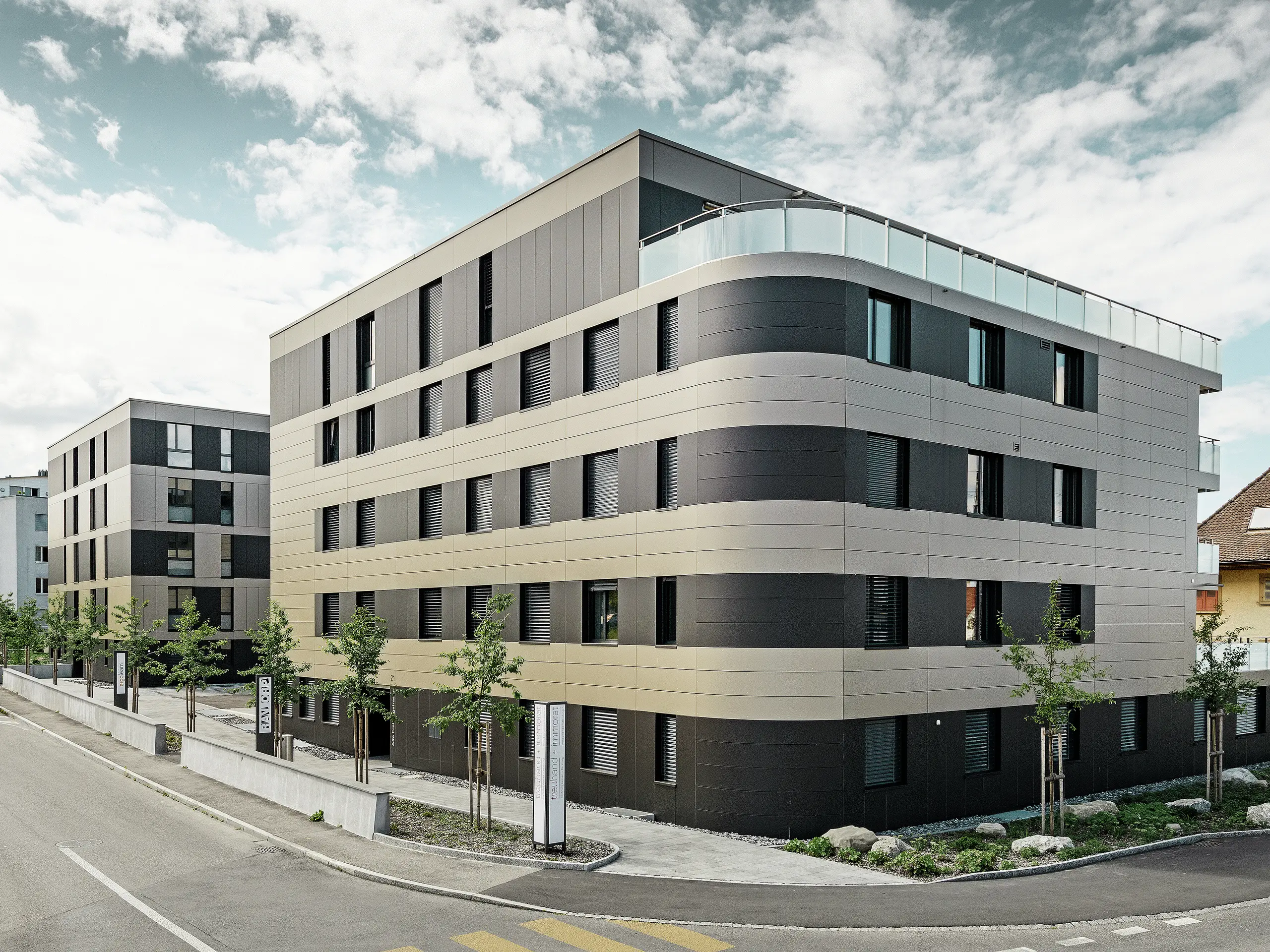 In der kubischen Wohnanlage von Murten (CH) sorgen die PREFA Aluminium Verbundplatten in Bronze und Schwarzgrau für eine optische Aufwertung und gleichzeitig für eine robuste und langlebige Fassade. Die Kombination dieser Farbtöne bietet ästhetische Akzente und schützt das Gebäude langfristig vor Wettereinflüssen. An der Gebäudeecke zur Straße hin ist eine Rundung zu erkennen, was das Rollbiegen der PREFABOND Aluminium Verbundplatten demonstrieren soll.