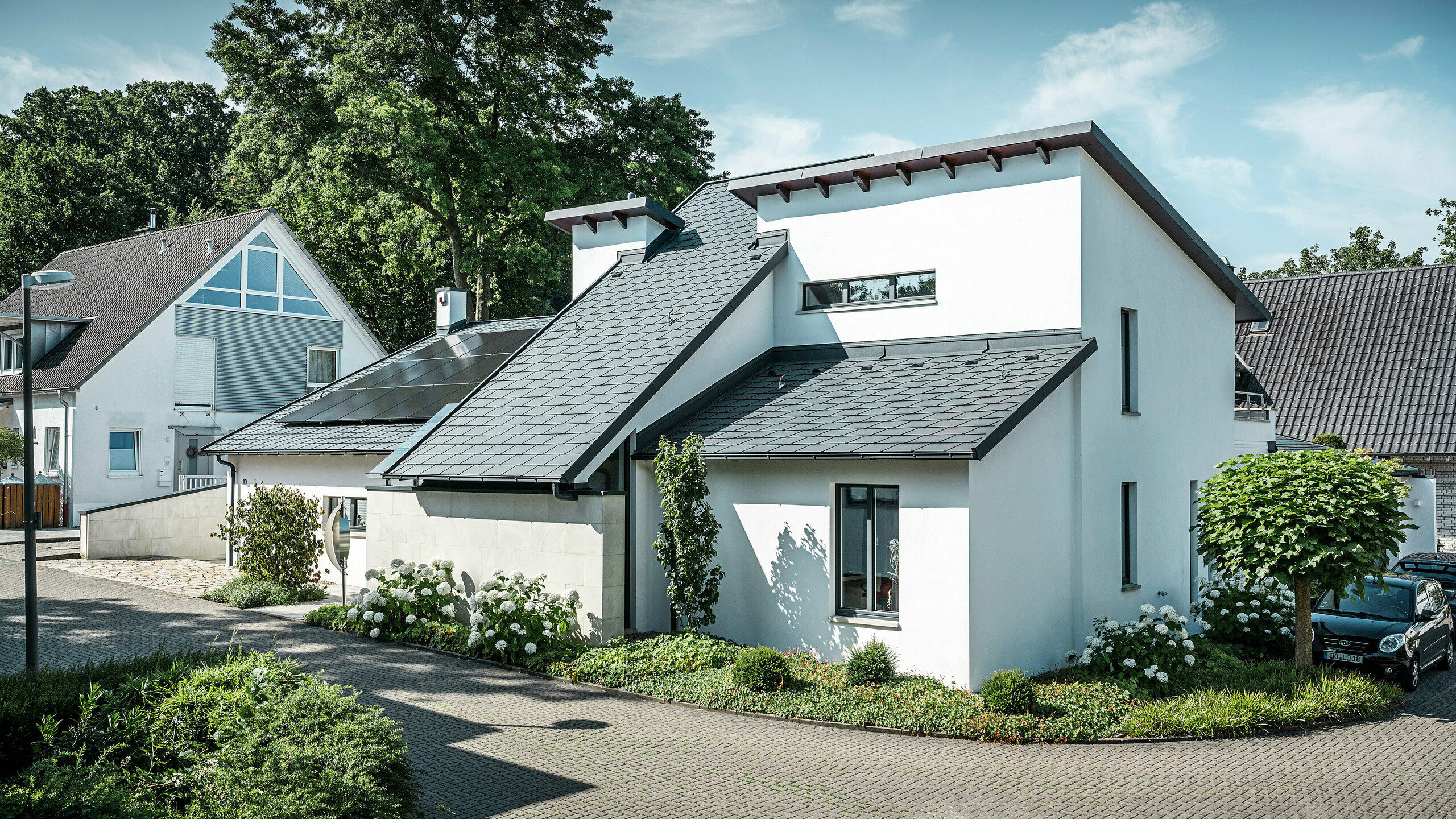 Modernes Einfamilienhaus mit weißer Putzfassade und dunklem PREFA Schrägdach aus Aluminium. Auf dem Dach sind integrierte Photovoltaikmodule und eine Aluminium-Dachentwässerung in Anthrazit zu sehen. Das Gebäude liegt in einer gepflegten Wohnsiedlung mit bepflanzten Vorgärten, gepflasterter Zufahrt und umliegenden Einfamilienhäusern.
