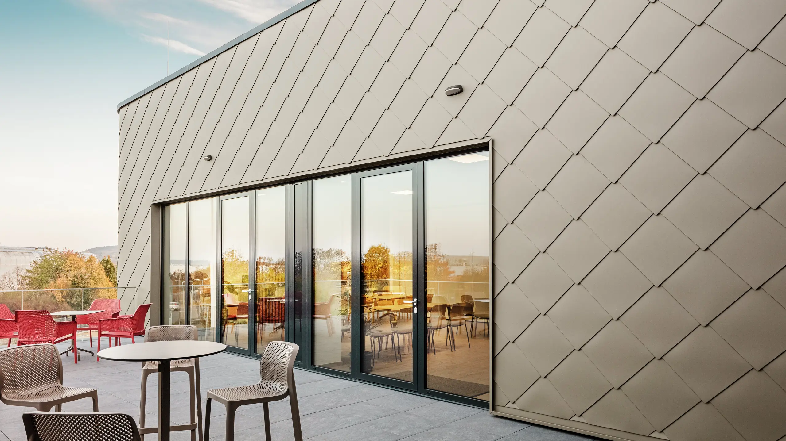 [Translate to Română:] Die Fassade des neuen PREFA Standortes in Törökbálint besticht durch die eleganten PREFA Wandrauten 44 × 44 in Bronze. Die präzise verlegte Aluminiumfassade sorgt für ein modernes, dynamisches Erscheinungsbild und bietet gleichzeitig maximalen Schutz vor Witterungseinflüssen. Die großzügige Glasfront verbindet Innen- und Außenbereich und schafft auf der großzügigen Terrasse eine helle und einladende Atmosphäre.
