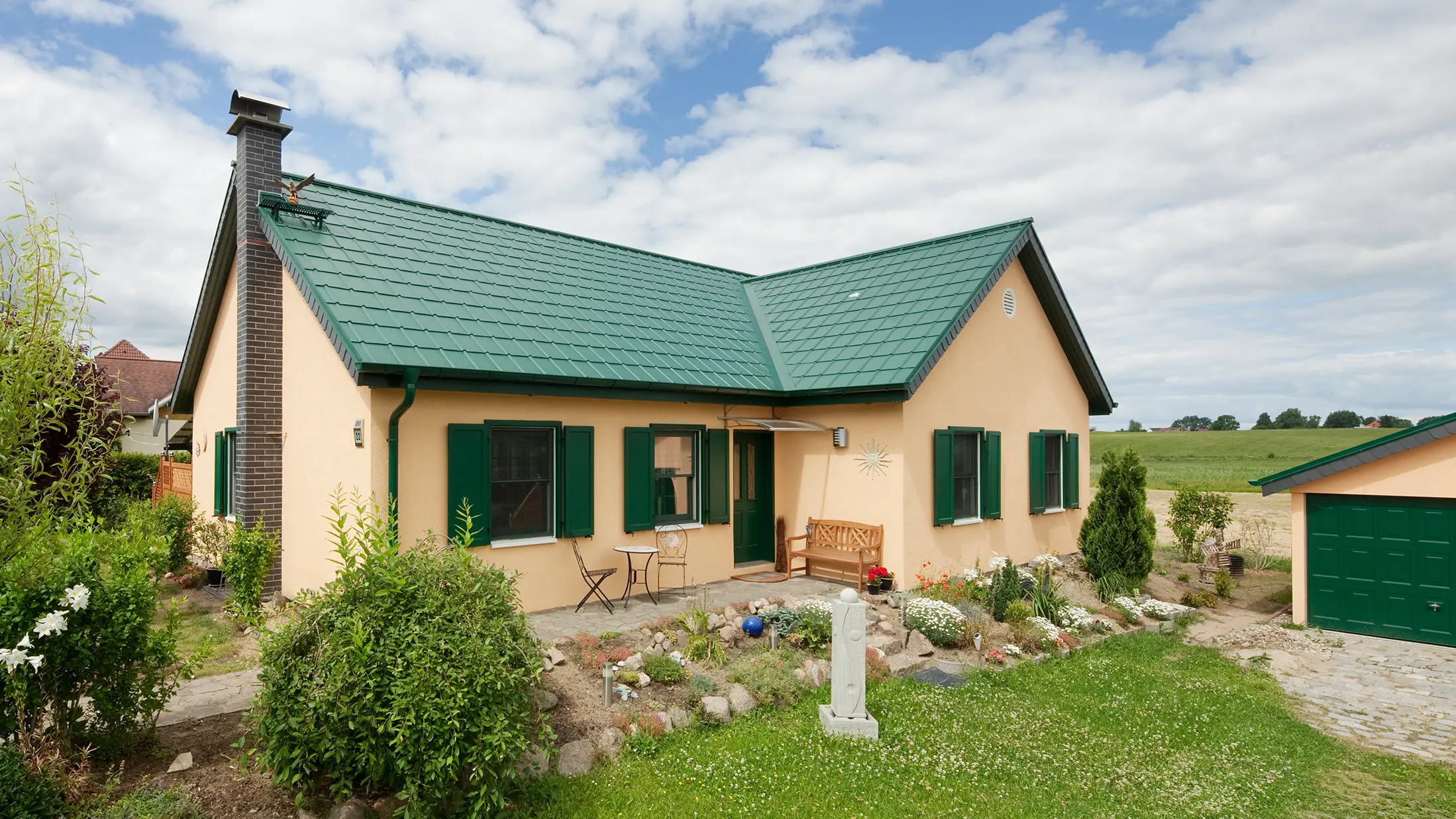 Încântătoarea vedere de ansamblu a casei monofamiliale din Friedrichswalde prezintă acoperișul durabil din aluminiu în verde mușchi, care împodobește atât casa de locuit, cât și garajul. Nuanța de verde a Plăcilor classic PREFA se integrează armonios în mediul natural și subliniază arhitectura simplă a casei. Acoperișul în două ape extins, în formă de L, se află în fața unui câmp. Căsuța cochetă are obloane verzi asortate și o mică grădină din față, foarte plăcută.