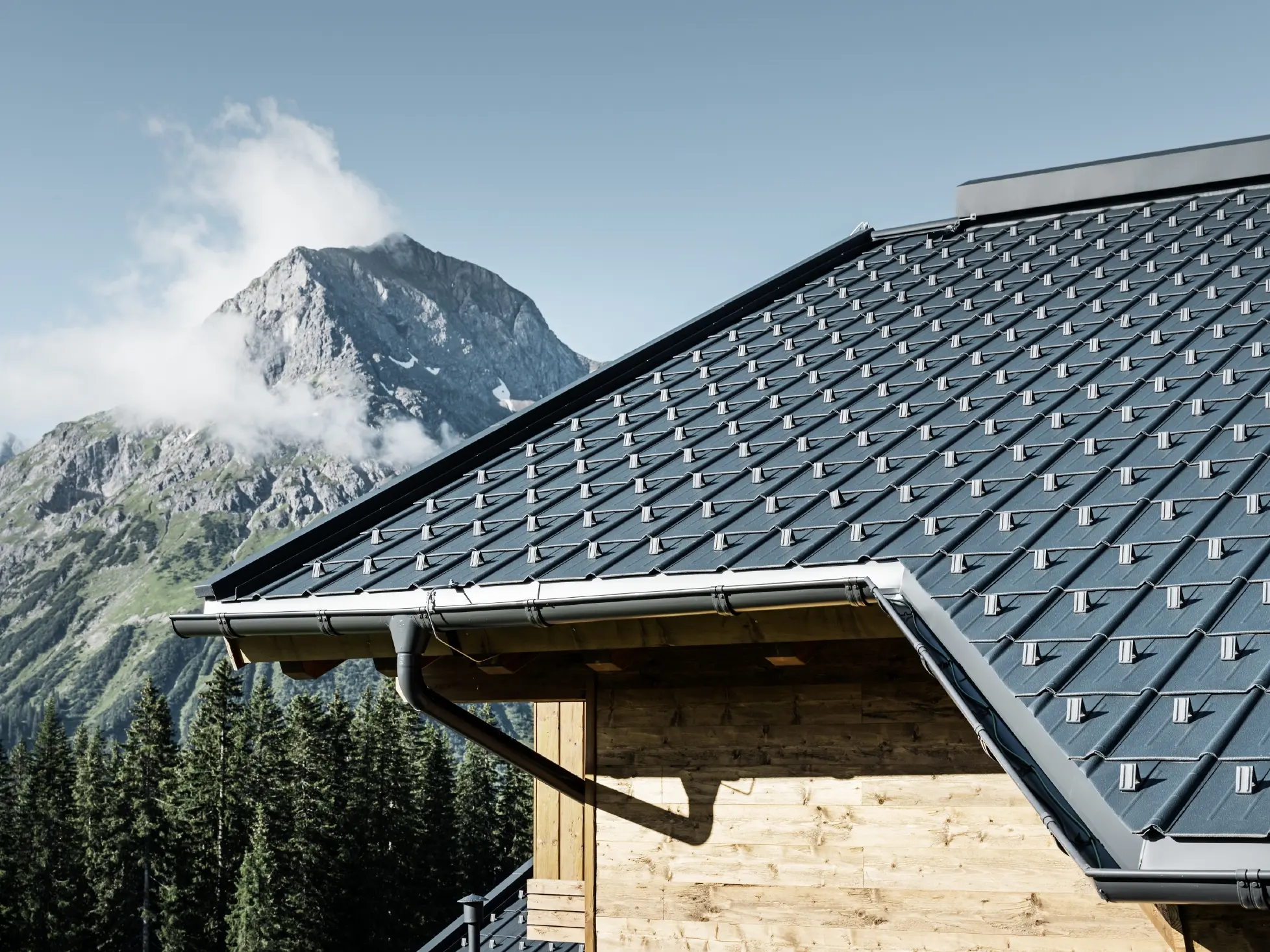 Placa classic PREFA în Lech am Arlberg, utilizare montană - placa classic în culoarea antracit instalată pe cabana din Lech am Arlberg, cu munți în fundal; jgheab PREFA în culoarea antracit; fațadă din lemn
