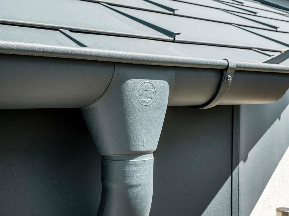 Prim-plan al acoperișului unei case cu placă de acoperiș PREFA R.16 în culoarea antracit și vedere detaliată a jgheabului și burlanului PREFA din aluminiu, asortate cromatic, pe timp însorit.