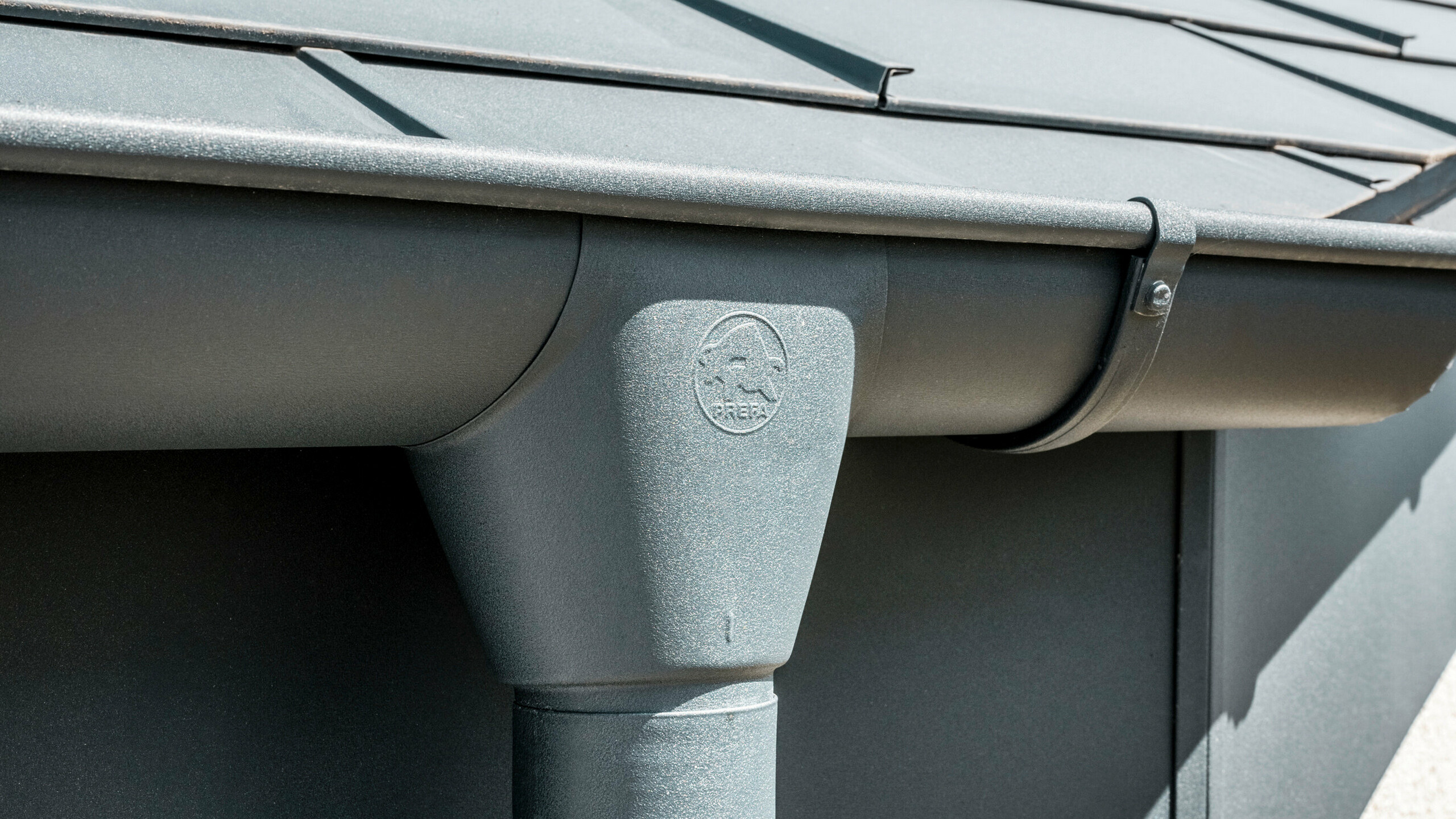 Prim-plan al acoperișului unei case cu placă de acoperiș PREFA R.16 în culoarea antracit și vedere detaliată a jgheabului și burlanului PREFA din aluminiu, asortate cromatic, pe timp însorit.