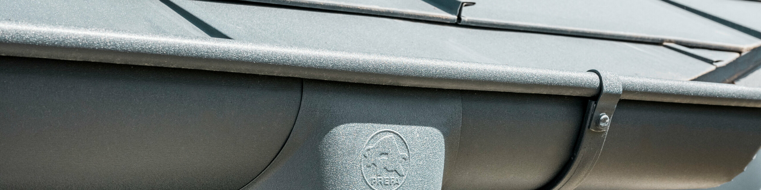 Prim-plan al acoperișului unei case cu placa classic PREFA R.16 în culoarea antracit, precum și vedere detaliată a jgheabului și burlanului PREFA din aluminiu, asortate cromatic, pe vreme însorită.
